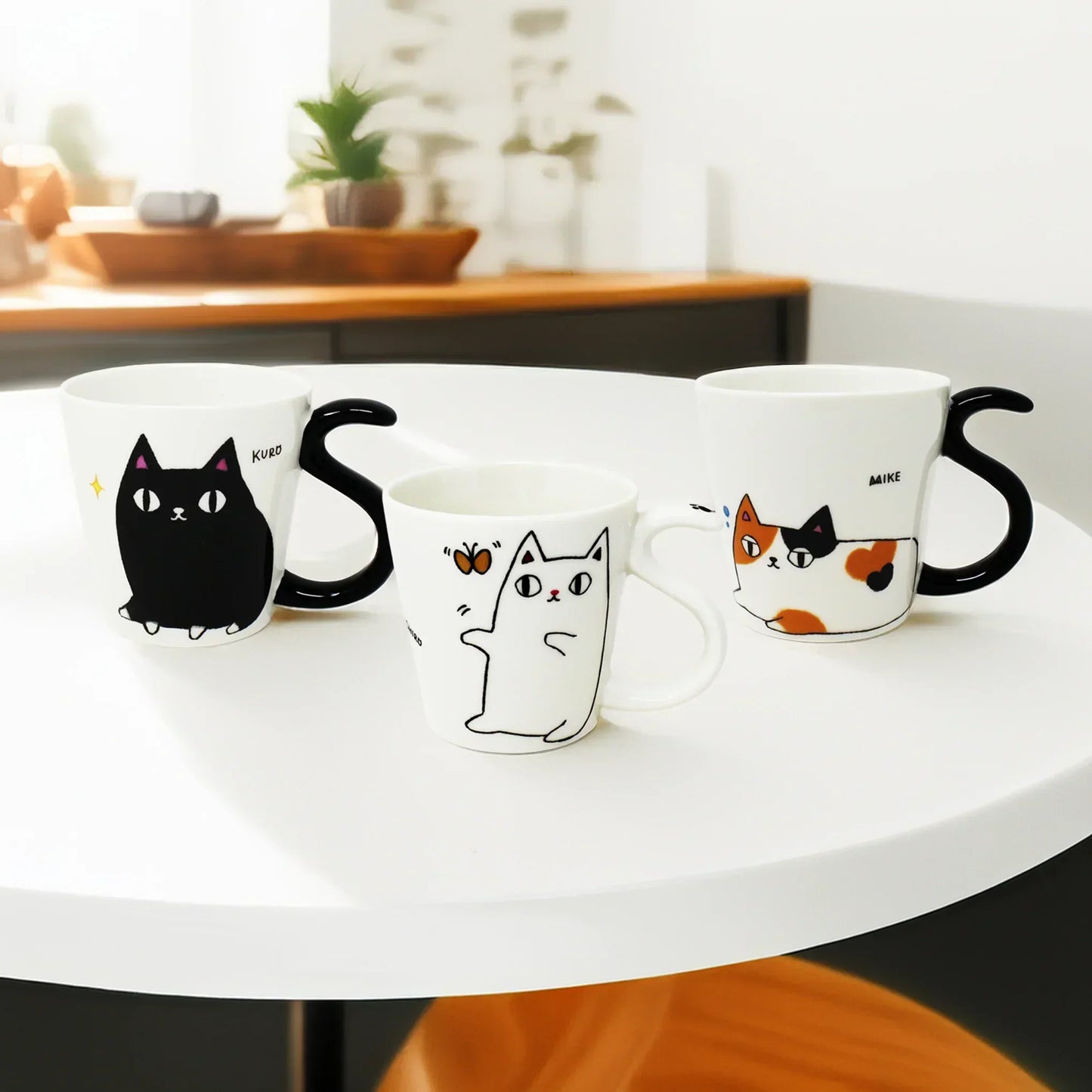 Three Cat Brothers porcelain mugs from Japan featuring Mike, Shiro, and Kuro with tail-shaped handles, displayed on a white modern dining table with a wooden chair, set against a kitchen counter background with a potted plant.