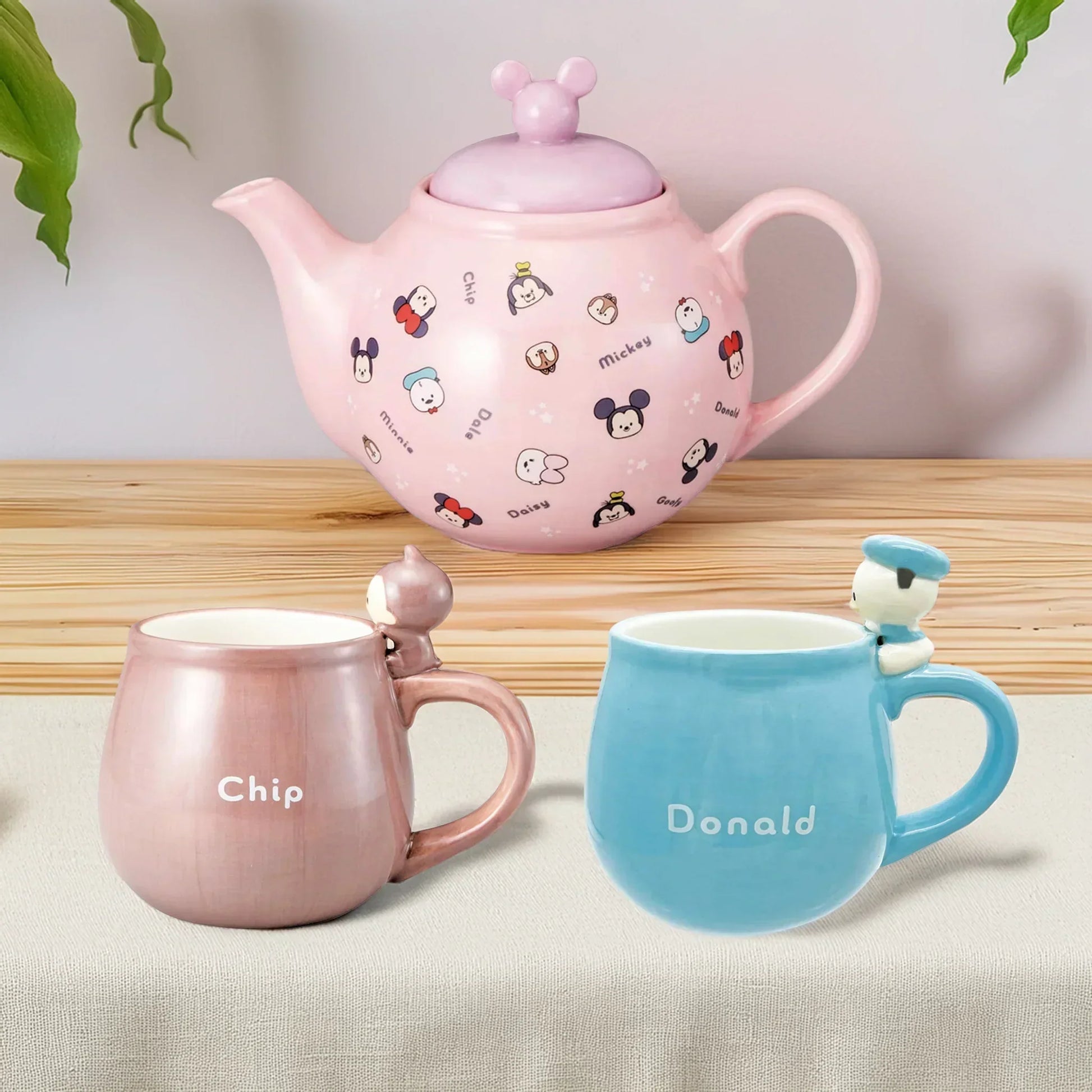 Pink teapot with Disney character designs, a pink mug labeled 'Chip', and a blue mug labeled 'Donald' on a wooden surface.