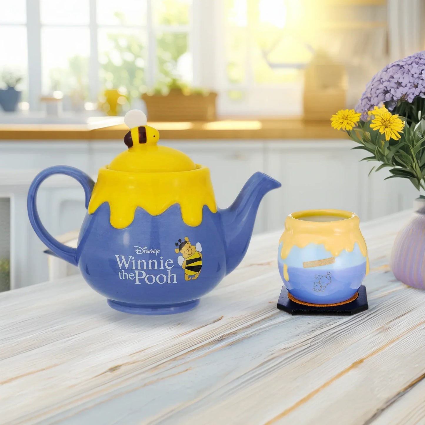 Winnie the Pooh-themed teapot and teacup on a kitchen counter with a window and flowers in the background.
