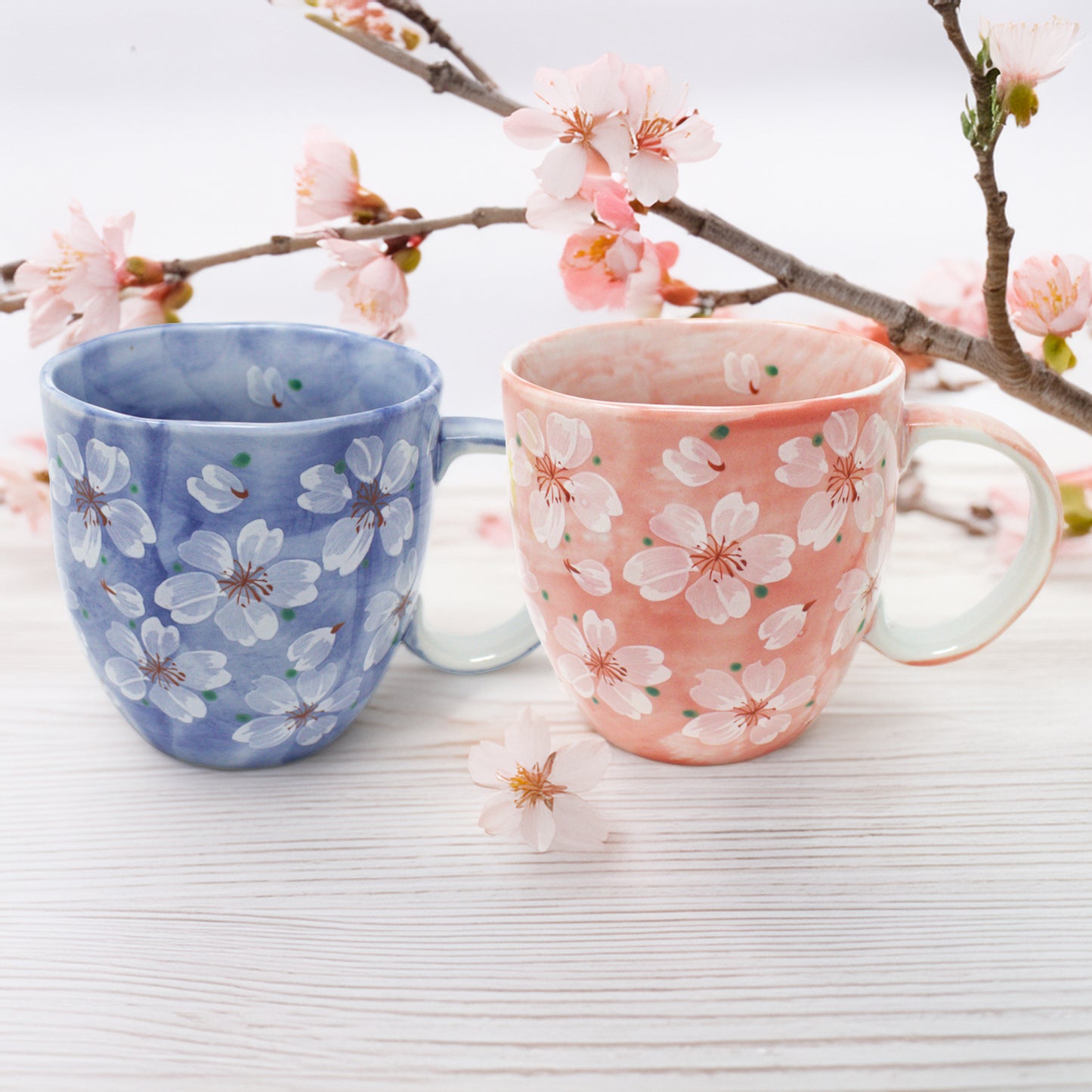 Two handcrafted Cherry Blossom Mino Ware ceramic mugs, blue on the left and pink on the right, featuring delicate sakura floral designs. Set on a realistic natural wood surface with subtle cherry blossom branches in a minimalist Japandi-style background. Soft natural lighting highlights the elegant porcelain finish.