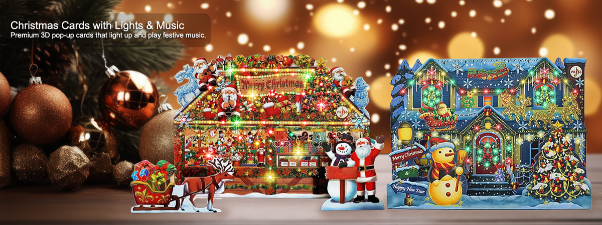 A festive Christmas card set displayed on a tabletop with holiday decorations. The set includes a large house-shaped card covered in colorful lights and ornaments, a smaller Santa-themed card with a snowman and sleigh, and a blue-toned holiday village card with glowing lights and a Christmas tree.