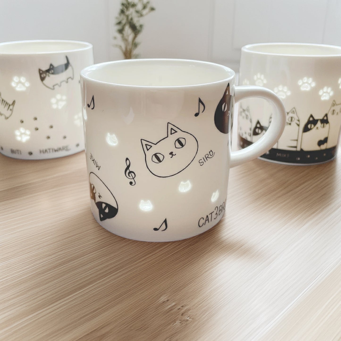 Three white mugs with black cat illustrations on a wooden surface.
