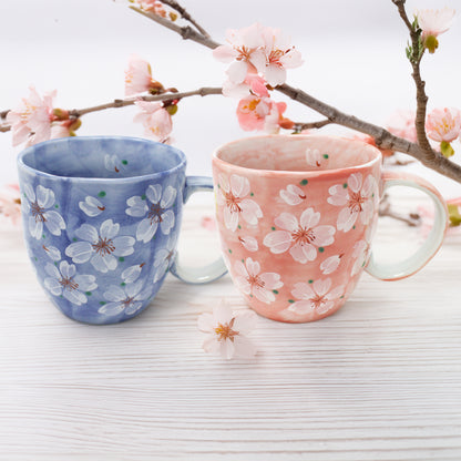 Two handcrafted Cherry Blossom Mino Ware ceramic mugs, blue on the left and pink on the right, featuring delicate sakura floral designs. Set on a realistic natural wood surface with subtle cherry blossom branches in a minimalist Japandi-style background. Soft natural lighting highlights the elegant porcelain finish.